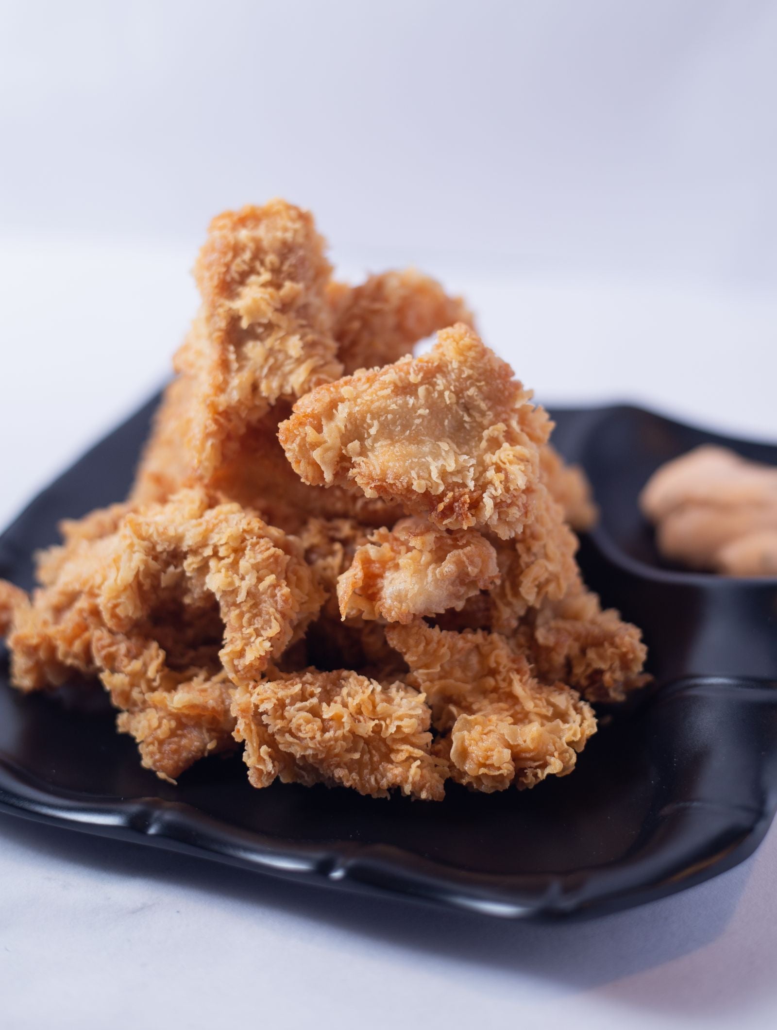 Stack of pale-gold broasted fried chicken strips with a thick craggy crust, piled on a matte black plate against a clean light grey background