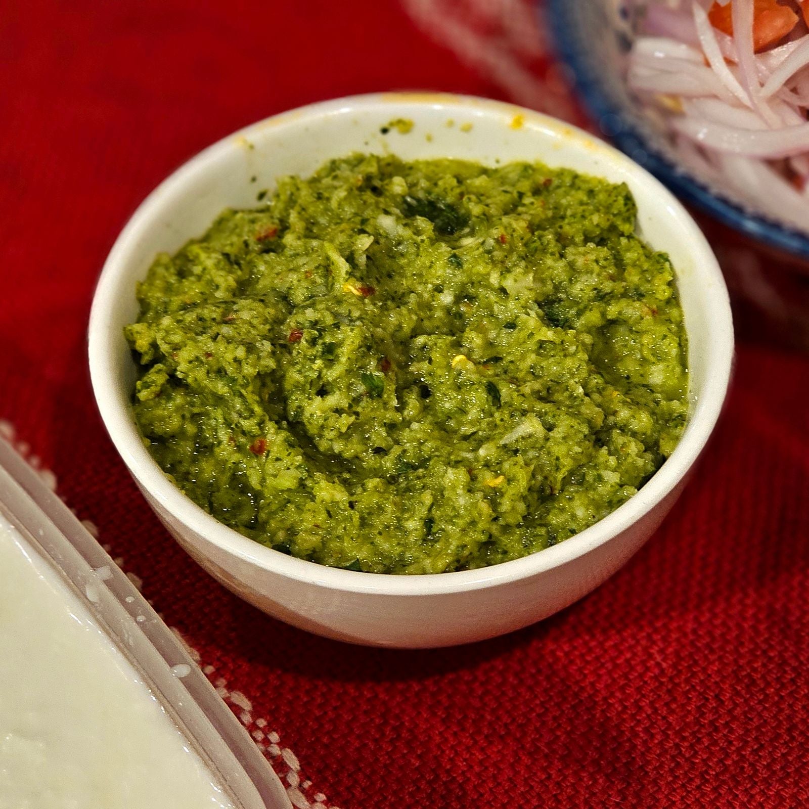 Vibrant green mint and coriander chutney with visible chilli flecks in a small white bowl on a red woven cloth