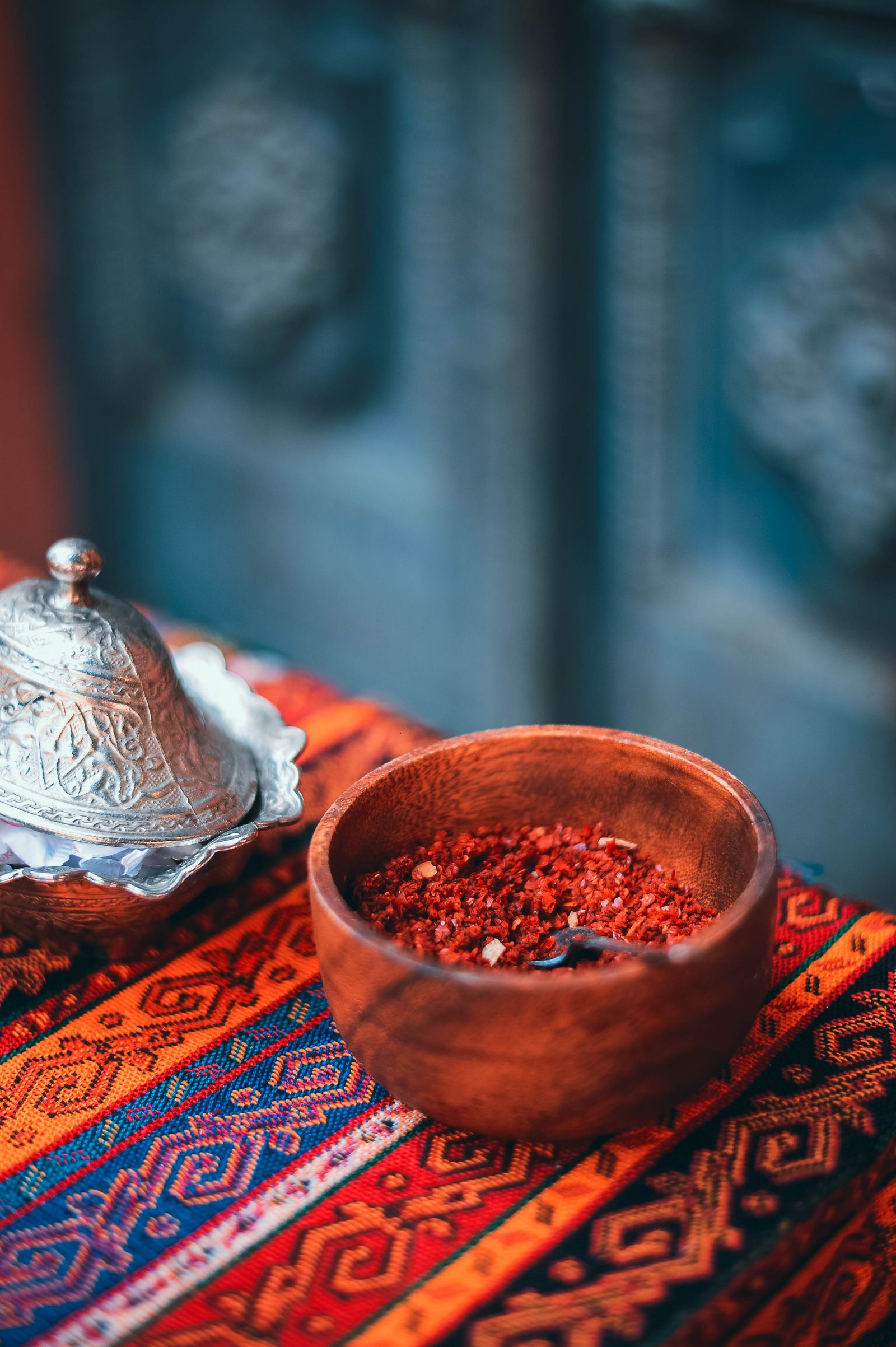 A rustic wooden bowl filled with coarse red sumac-style spice blend, resting on a vibrant patterned woven cloth beside an ornate embossed silver lidded vessel