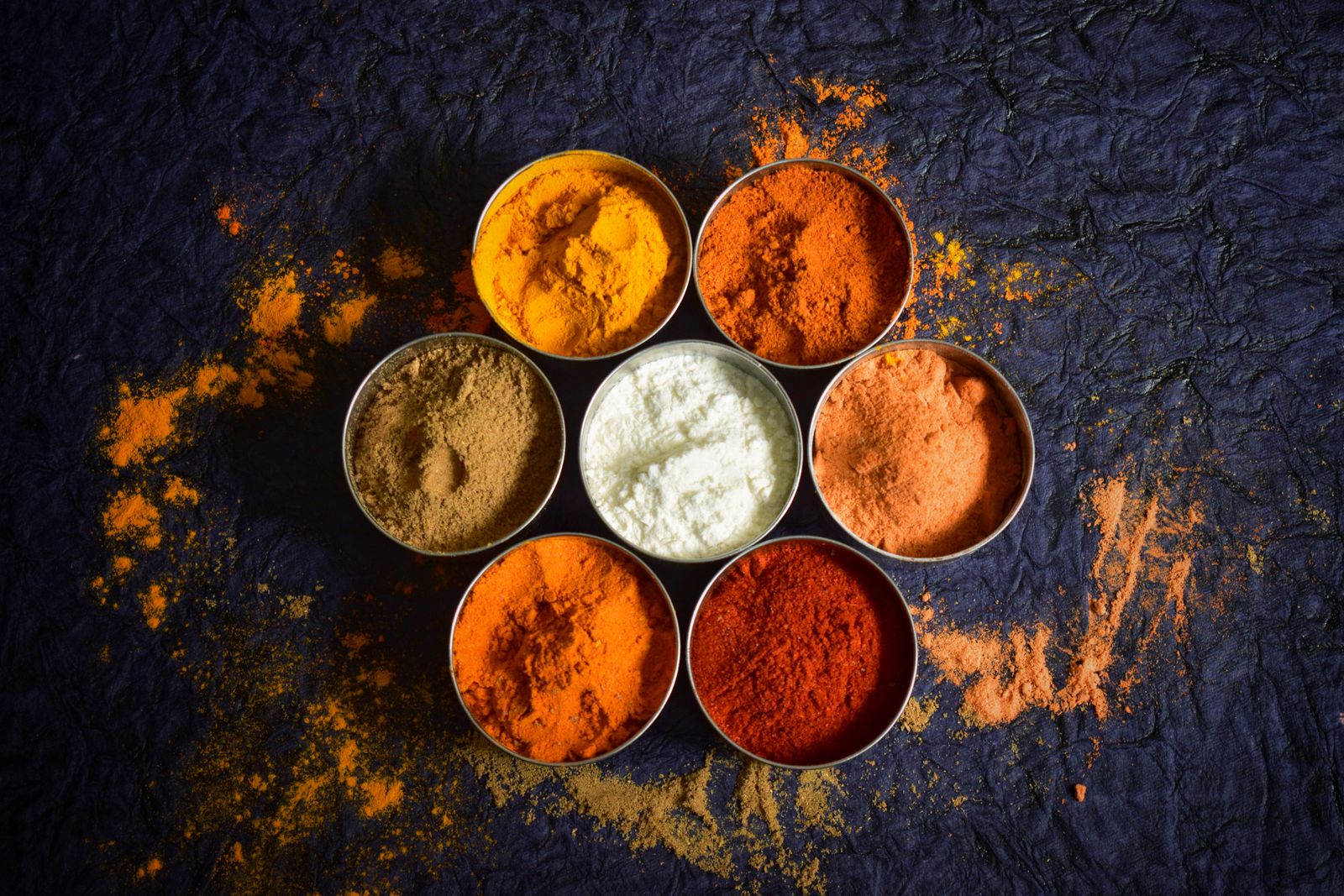 Seven metal bowls of colourful Indian spice powders — turmeric, chilli, paprika and ground masalas — arranged in a flower pattern on a dark textured surface with powder scattered around them