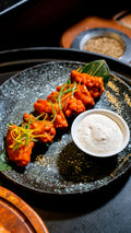 Bright orange buffalo-sauced chicken wings arranged on a dark speckled ceramic plate with a small white bowl of creamy dip and fine strips of spring onion and chilli