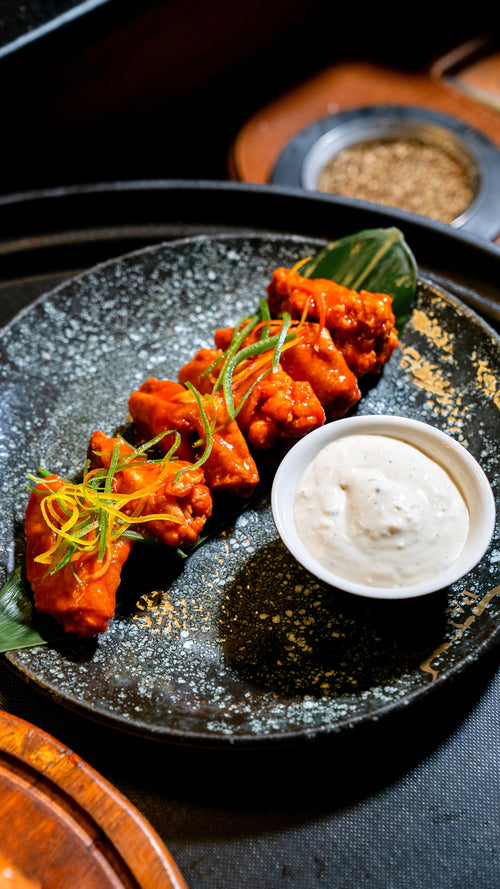Bright orange buffalo-sauced chicken wings arranged on a dark speckled ceramic plate with a small white bowl of creamy dip and fine strips of spring onion and chilli