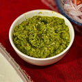 Vibrant green mint and coriander chutney with visible chilli flecks in a small white bowl on a red woven cloth