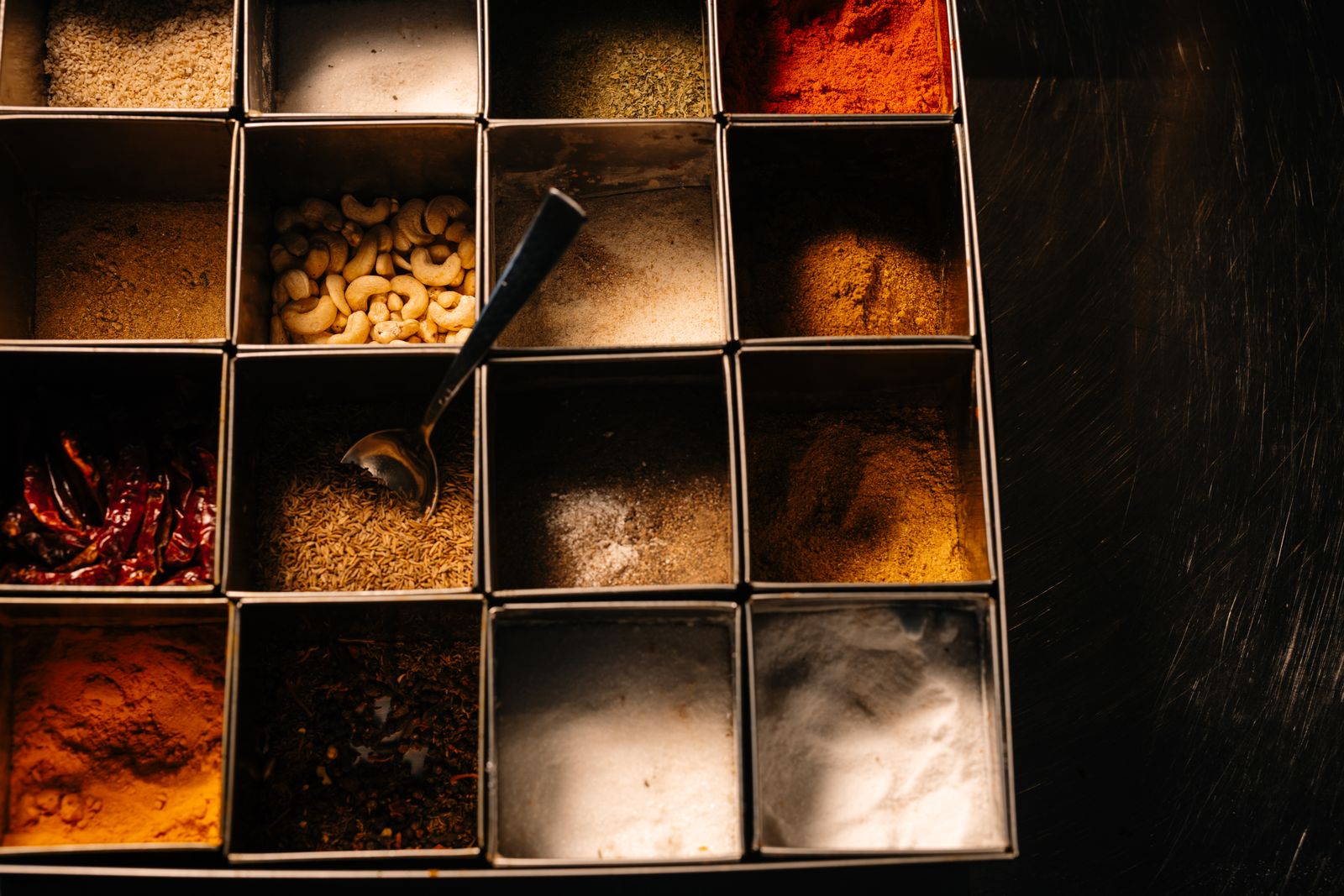 Overhead view of a compartmented metal spice box holding sixteen different spices — paprika, turmeric, chilli, cumin seeds, salt and cashews — with a small spoon resting in one compartment, on a dark wooden surface