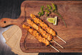 Three grilled marinated chicken skewers on a rustic dark wooden board with citrus wedges and fresh herbs