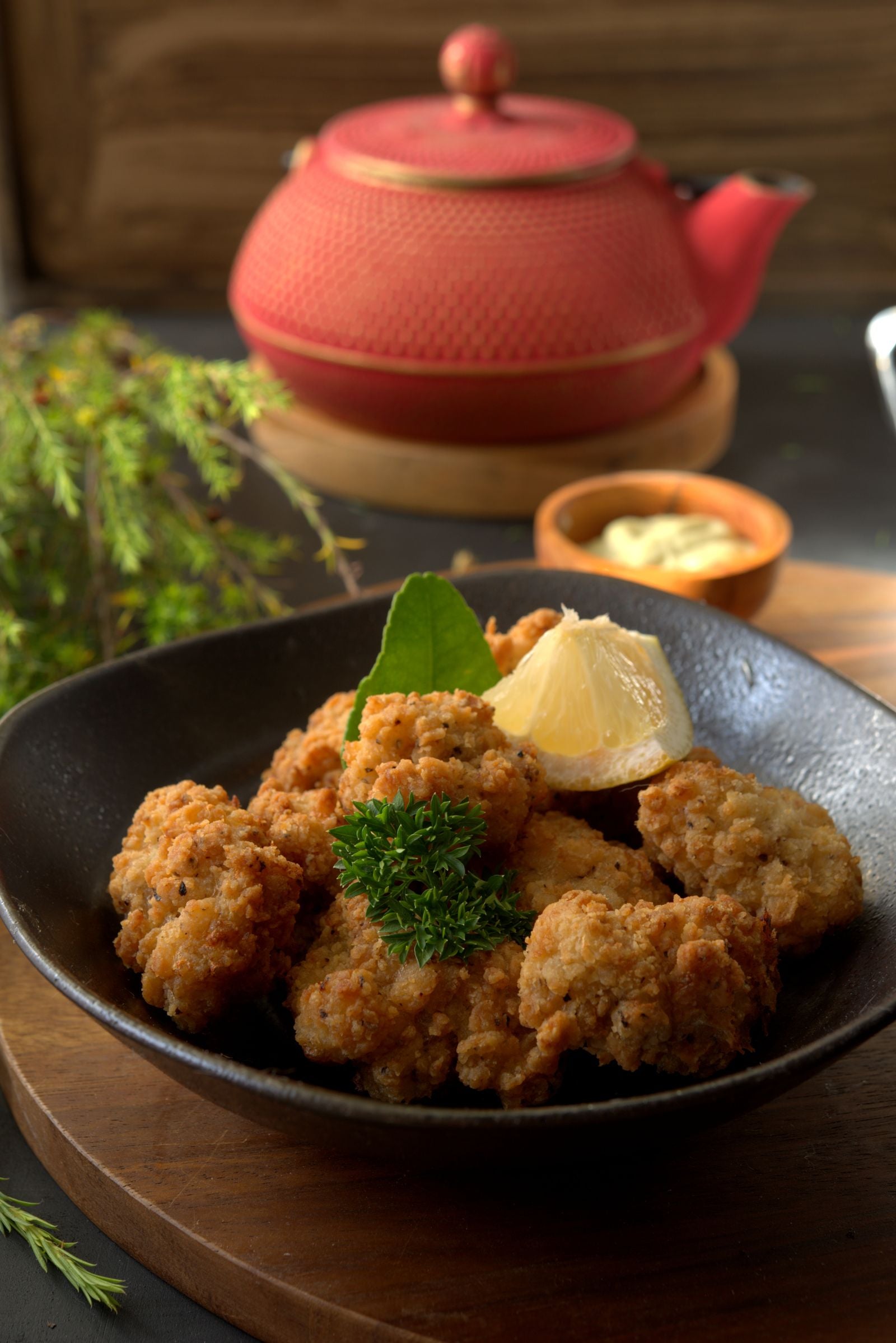 Craggy golden-crusted fried chicken pieces in a matte black bowl with a lemon wedge and a sprig of fresh green herbs, on dark wood