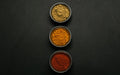 Three round metal bowls of spice rub powders — ground brown seasoning, orange paprika and deep red paprika — lined up vertically on a matte black surface