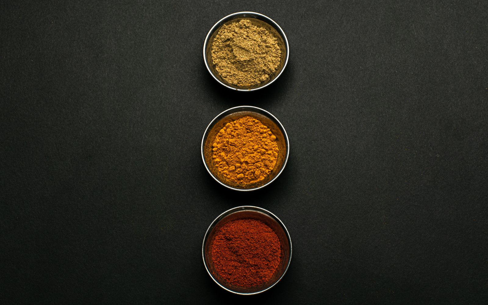 Three round metal bowls of spice rub powders — ground brown seasoning, orange paprika and deep red paprika — lined up vertically on a matte black surface