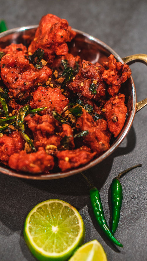 Crispy deep-fried chicken pieces in a bright red masala crust, tossed with fried curry leaves and garlic, served in a copper kadai on a dark surface with lime and green chillies