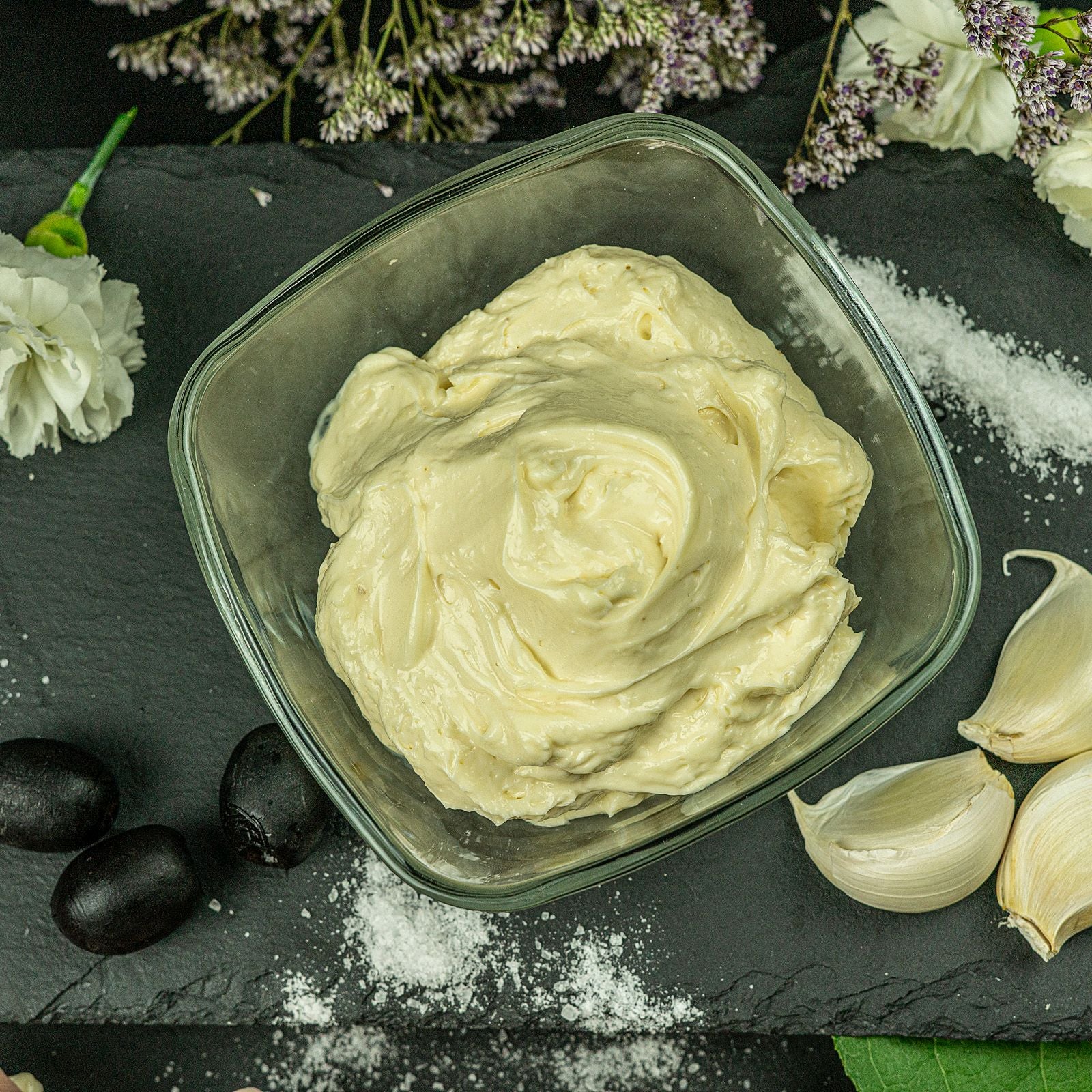 Thick whipped white garlic dip in a square glass bowl on black slate, surrounded by peeled garlic cloves, coarse salt and black olives