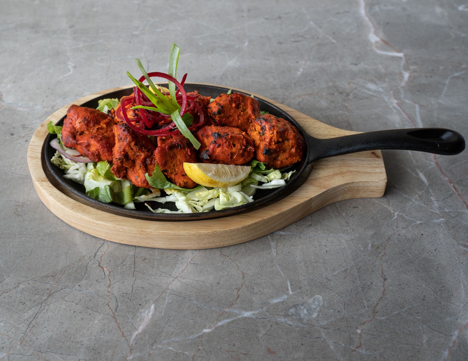 Charred red tandoori chicken pieces on a black cast-iron sizzler plate with shredded cabbage, pickled onion and a lemon wedge